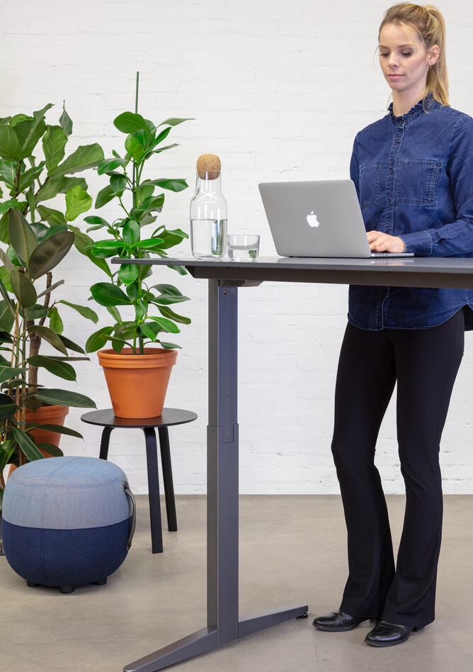 Balance Lift, Recharge 45 Royal Ahrend Balance Lift workstation in grey with charcoal top in stand position and blue Recharge 25 stool with female model standing front right detail view